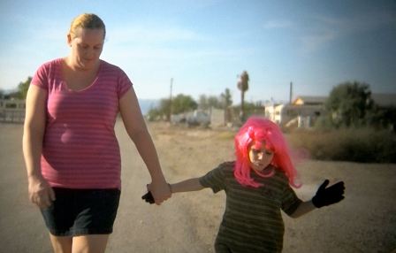 Filmstill aus "Bombay Beach": Mutter spaziert mit Kind, das eine rosa Perücke trägt, in öder Landschaft